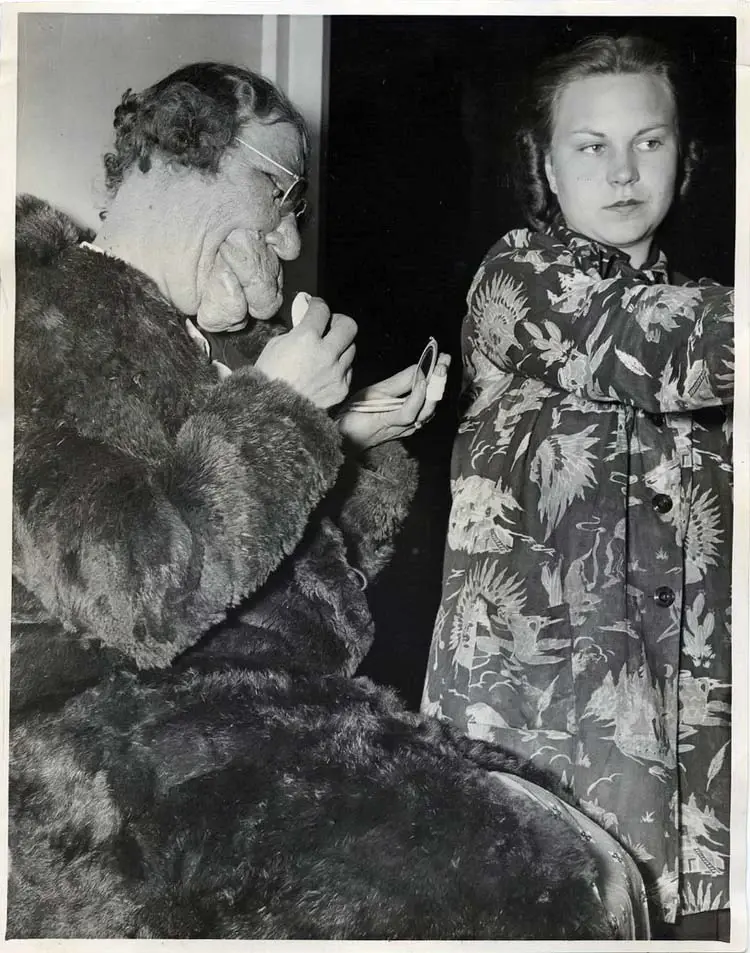 Older person in fur coat examines paper; young woman stands beside, wearing a patterned robe, appearing focused. Dimly lit setting.