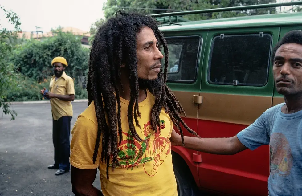 Three men stand in front of a parked van. The man in the foreground wears a yellow T-shirt with vibrant patterns. Another observes from behind.