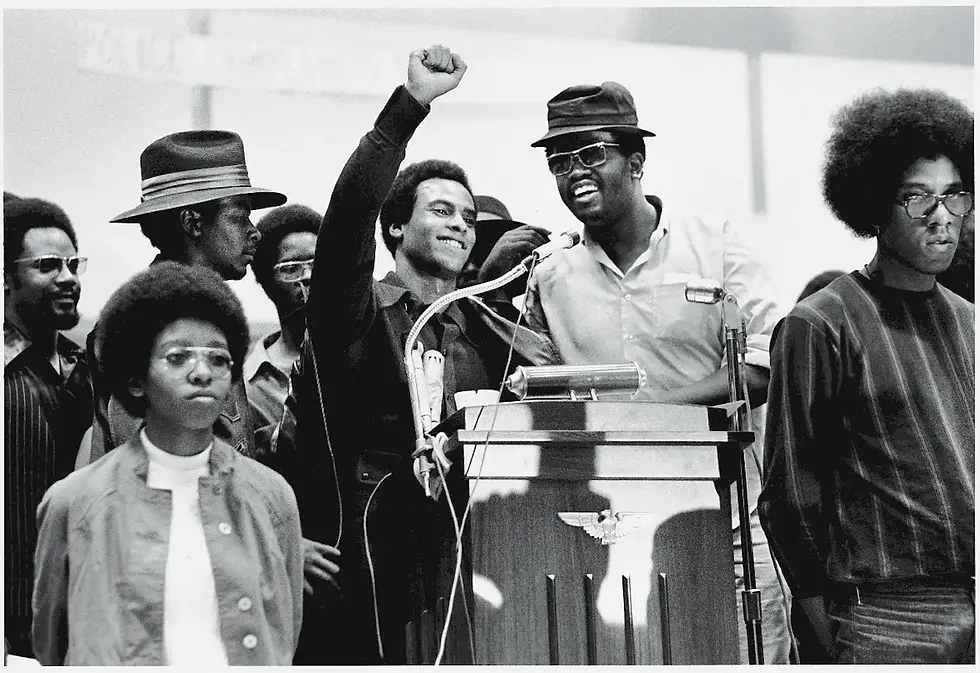 Group of people at a podium; one raises a fist. Others stand nearby, serious. Black and white photo conveying unity and determination.