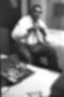 Man in a white shirt sits on a bed, singing passionately, holding a cord. A LIFE magazine is visible on the bed. The room is dimly lit.