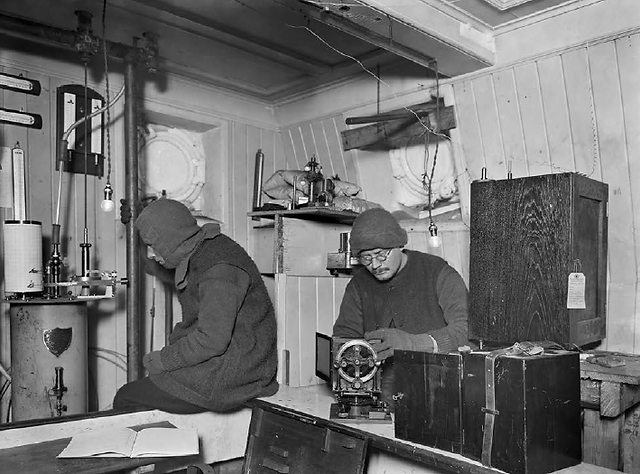 Two men operating scientific equipment within a ship's compact cabin.