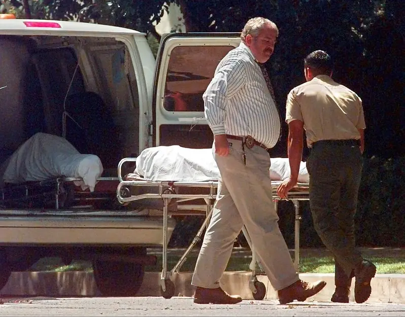 Two men move a stretcher with a covered body towards a van in a sunny outdoor setting. One wears a striped shirt, the other in beige.