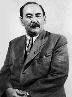 Man in a suit sitting against a plain background. He wears glasses and has a mustache, conveying a serious expression.