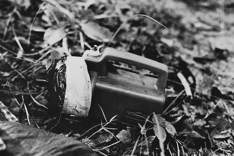 A black-and-white photo of a portable cassette player on the ground, surrounded by leaves and grass, creating an abandoned, nostalgic mood.