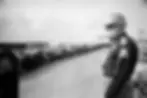 People kneeling in a long line on a street, overseen by a state trooper in uniform. Background shows buildings with visible signs. Black and white.