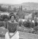 Smiling woman in a striped top and necklace sits on a ledge. Overlooks a scenic landscape with trees and hills. Black and white image.