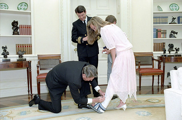 George H.W. Bush kneels, helping a young girl with her sandal.