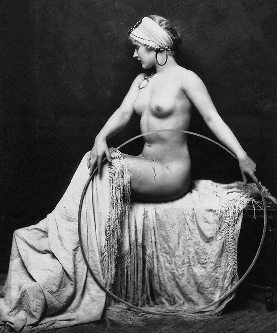 Nude showgirl seated side-on with a hoop and head wrap, posed against a dark background.
