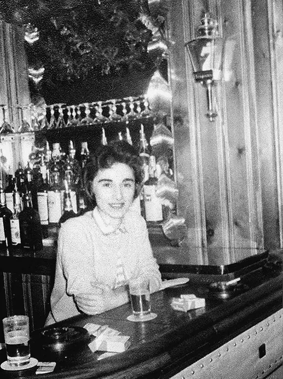 Woman smiling behind a bar with bottles and glasses. Wood-paneled background, dim lighting, and two drinks with ashtrays on the counter.