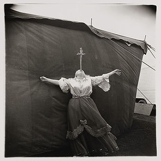 Woman with outstretched arms, cross at neck, against dark fabric wall.