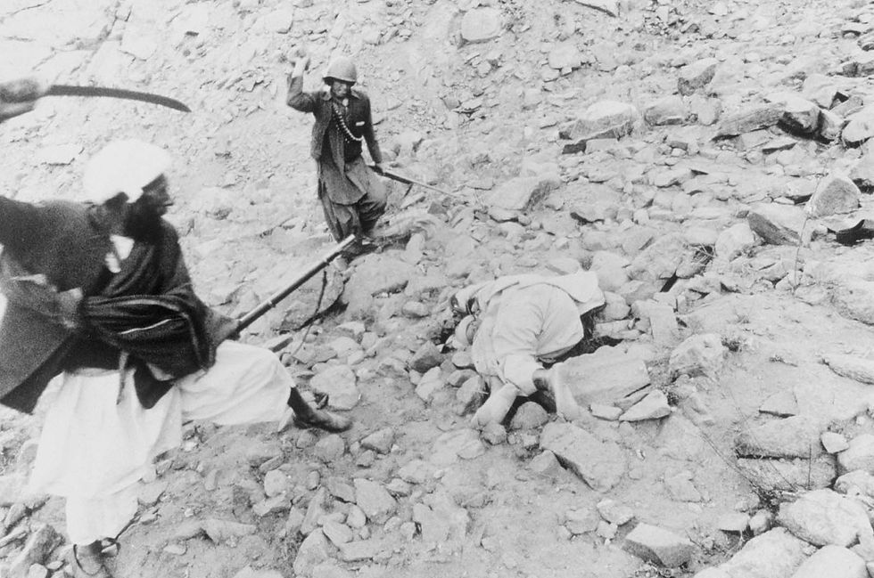 Two men, one with a rifle, another with a sword, attack a figure lying on rocky ground. The setting is rugged, evoking tension and conflict.