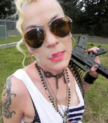 Chrese Evans with blonde hair and sunglasses holding a rifle over their shoulder. Wearing multiple necklaces in a grassy outdoor setting.