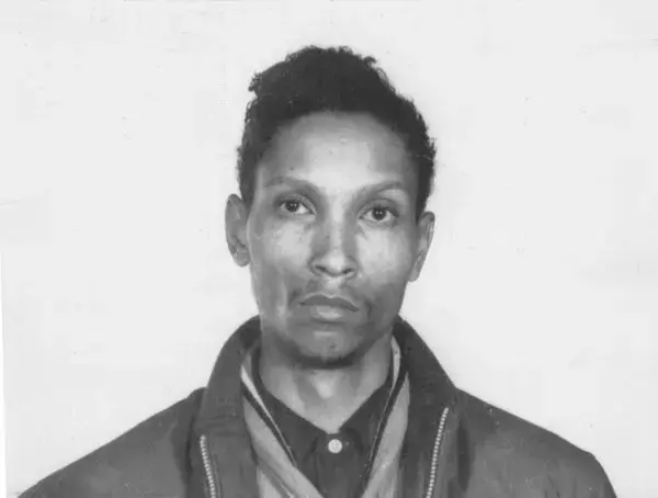 A black-and-white portrait of a person with short, curly hair, wearing a jacket and shirt. The background is plain. The expression is neutral.