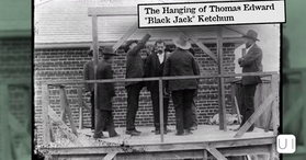 Men gather on a wooden gallows platform, adjusting a noose over a suited man, with a brick building behind.