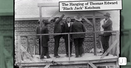 Men gather on a wooden gallows platform, adjusting a noose over a suited man, with a brick building behind.
