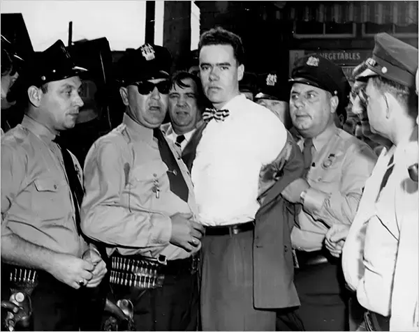 A group of police officers surrounds a man in a bow tie. The man appears calm. The background shows a building and a sign with "TABLES."