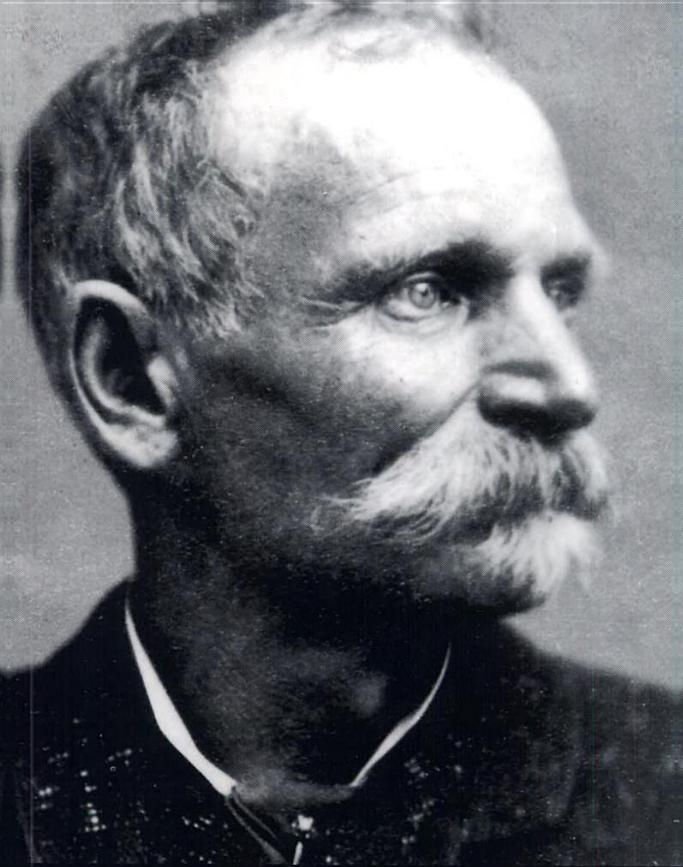 Elderly man with a large mustache gazes to the side in a black-and-white portrait. He has a serious expression, wearing a dark suit.