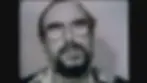A man with glasses and a beard looks upward. The grayscale image has a plain background. Mood appears serious. No text visible.