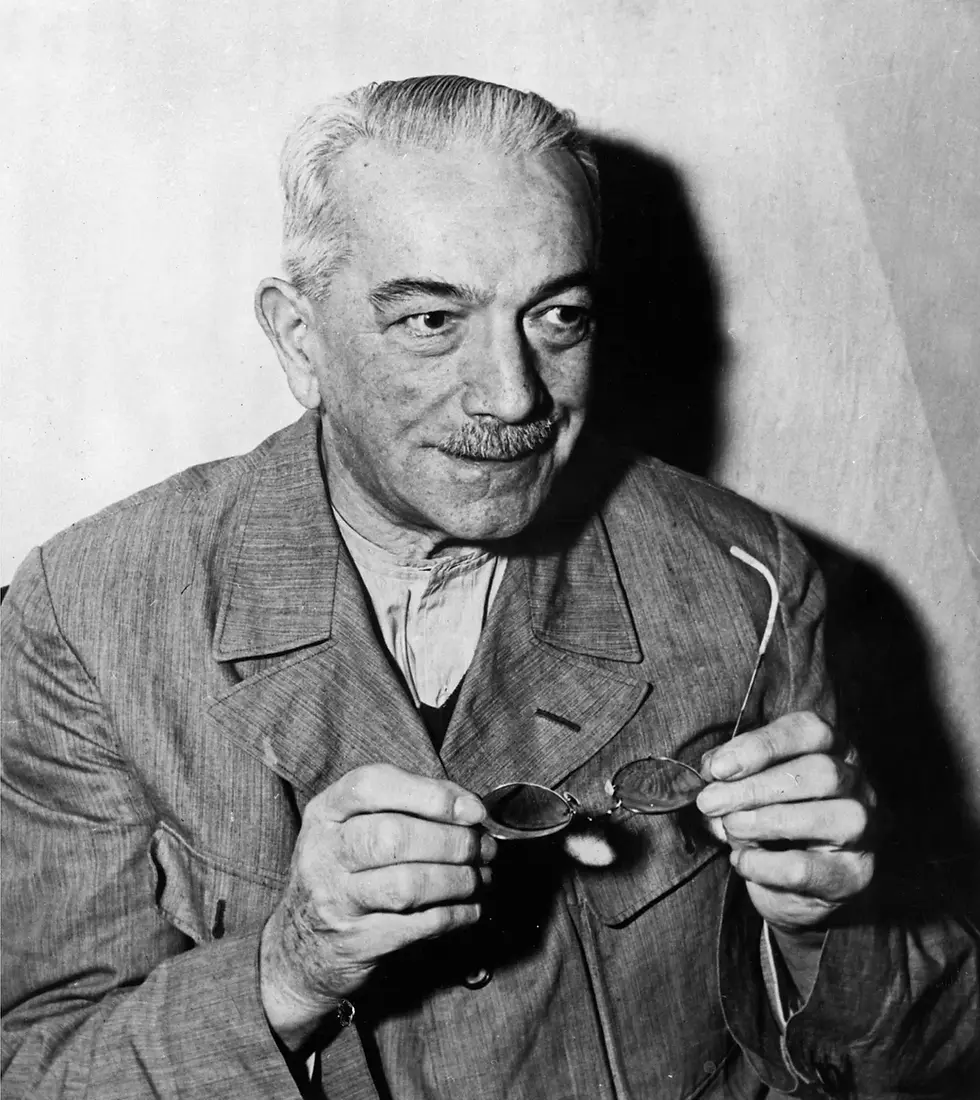 Elderly man with a mustache holds glasses, wearing a suit. Neutral expression. Monochrome background and attire.