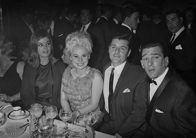 Four people in formal attire seated at a table with drinks and a "Schweppes" bottle, smiling and chatting in a crowded, elegant setting.