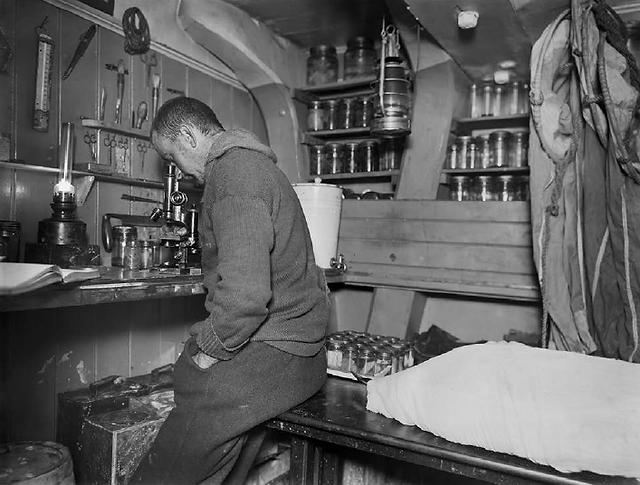 Man sits, head bowed, working at cluttered workbench in ship's interior.