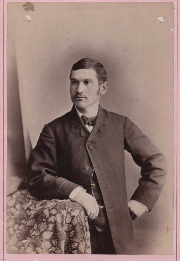 Man in a suit poses with hand on floral-patterned table in vintage sepia-tone setting, exuding a formal, composed mood.