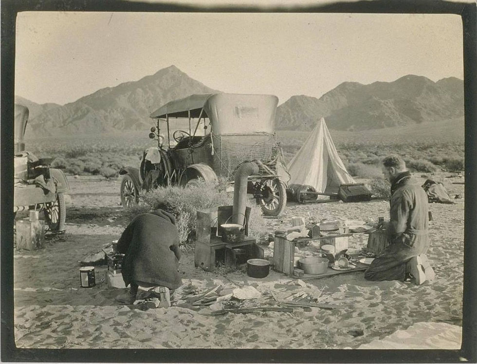 Desert travelers cooking beside vintage car and tent, distant mountains.