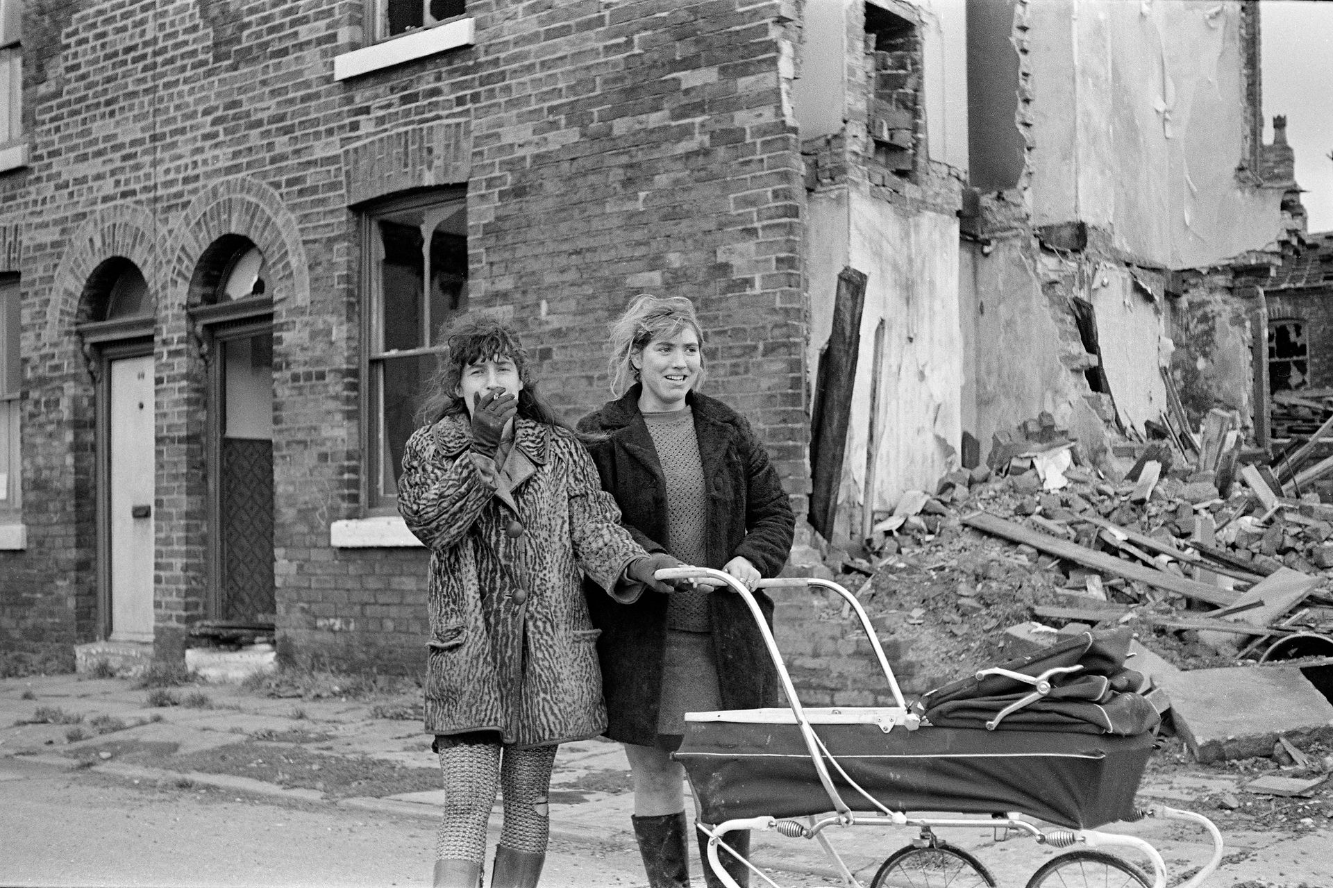 Two women pushing a baby carriage past demolished brick buildings.