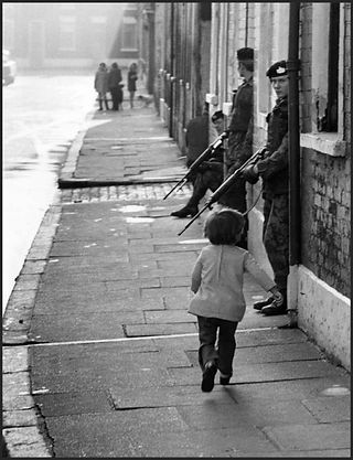 Small child walks past two armed soldiers on a city street.