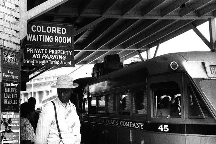 Man in white hat under "Colored Waiting Room" sign next to a bus. Nearby posters display text. The setting is monochrome and somber.