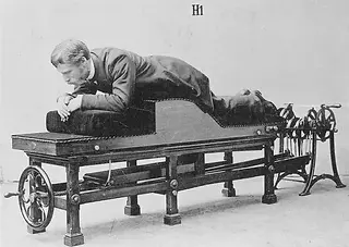 Man lying face down on antique mechanical exercise machine, visible text H1.