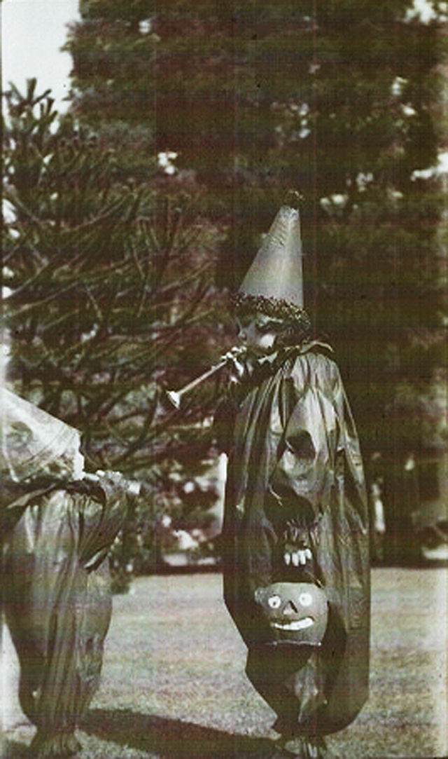 Child in Halloween costume with pointed hat, holding a jack-o'-lantern and playing a horn. Trees in the blurry background. Sepia tone.