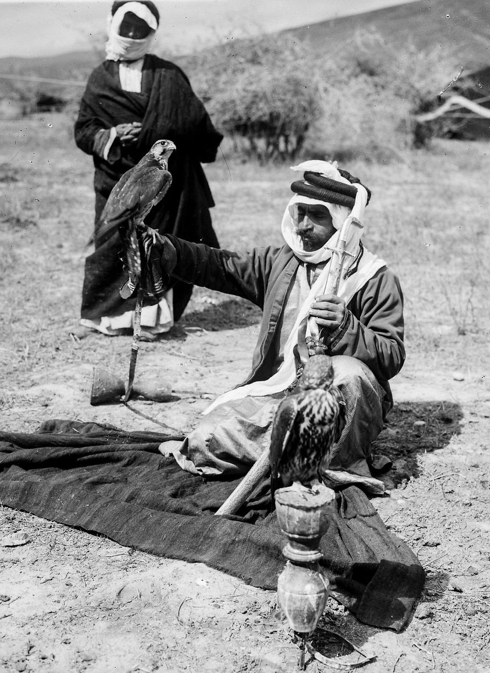 Black-and-white photograph of a Bedouin man seated on a blanket outdoors, holding a falcon on his gloved hand while another falcon rests on a perch in front of him. He wears traditional robes and a headscarf, with a musket resting across his lap. Captured in the early 20th century by the American Colony Photo Department.