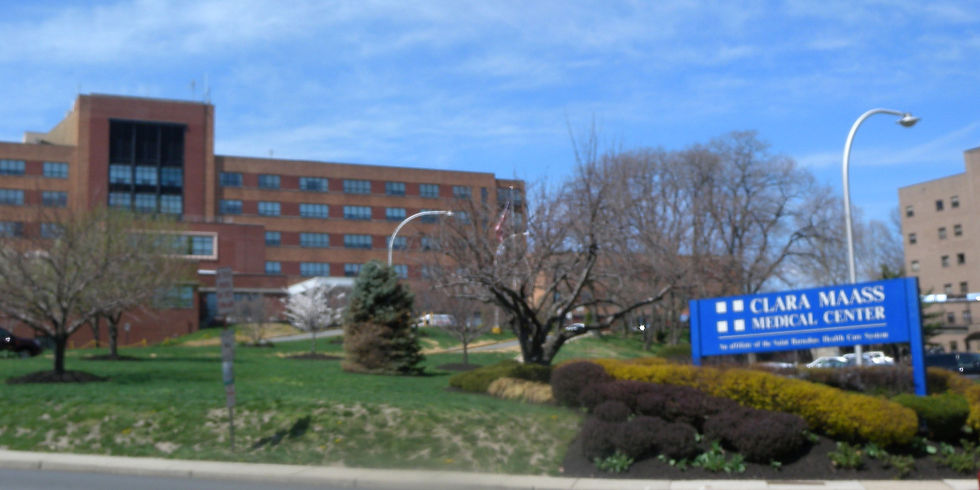 Brick building with trees and a blue sign reading "Clara Maass Medical Center" in the foreground. Clear sky and an urban setting.