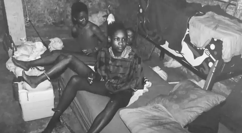 Three people are sitting in a dim room on disheveled bedding. One is in focus, wearing a patterned shirt. They appear somber.