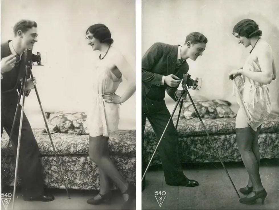 Man in suit takes photo with vintage camera of smiling woman in lace dress. Floral pillow in background. Sepia tone, cheerful mood.
