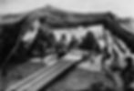 People sit under a makeshift tent in a desert setting, engaged in weaving on a loom. Monochrome tones, no visible text. Mood: serene.
