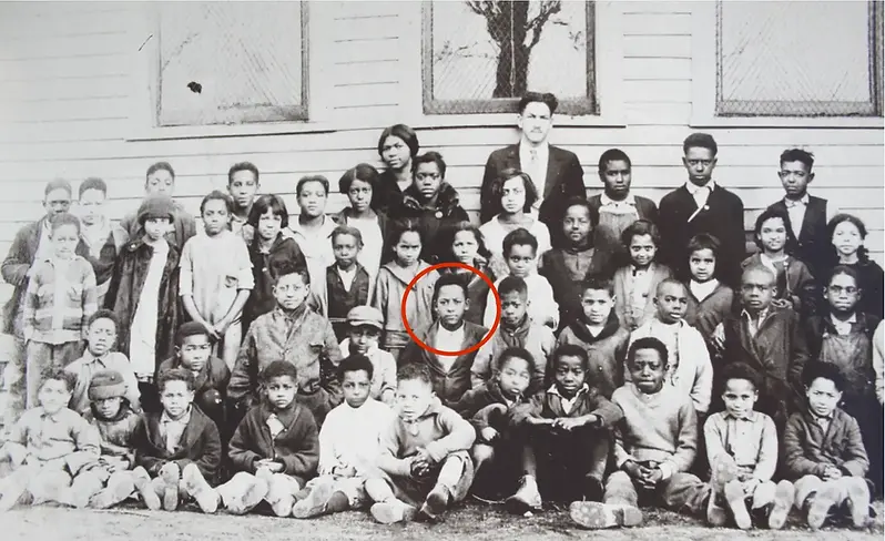A vintage black-and-white photo of a diverse group of schoolchildren and a teacher. One child is circled in red. They stand in front of a building.