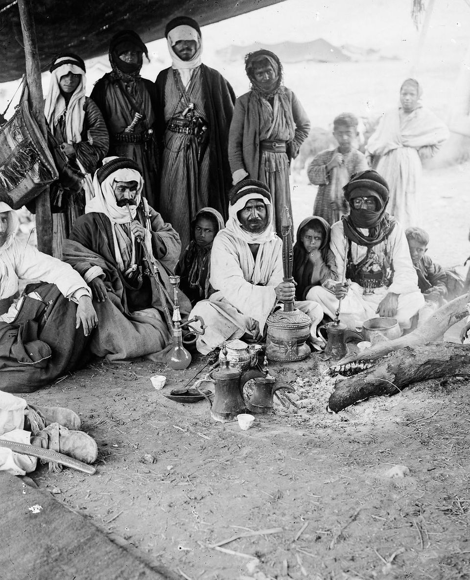 Black-and-white photograph of a group of Bedouin men and women gathered around a coffee set under a tent, with ornate pots and traditional hookahs placed on the ground. Some sit cross-legged while others stand behind them, dressed in layered robes and keffiyehs. Taken in the early 20th century by the American Colony Photo Department.
