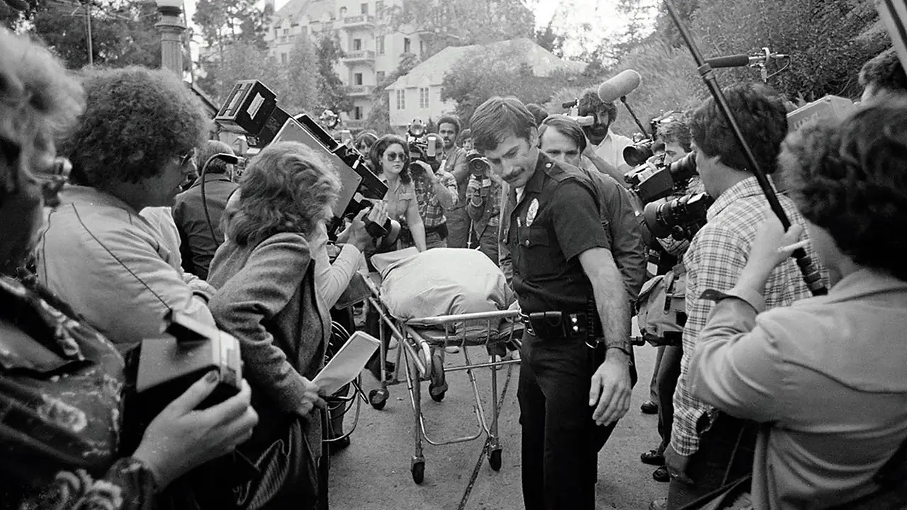 Police officer escorts covered body on stretcher, amidst cameras and onlookers.