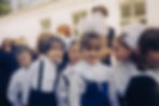 Children dressed in white and blue uniforms with lace, smiling in a school lineup outside a building. The mood is joyful and playful.