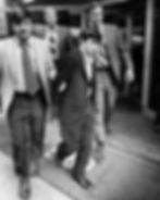 Young man in suit apprehended, escorted by plainclothes officers.
