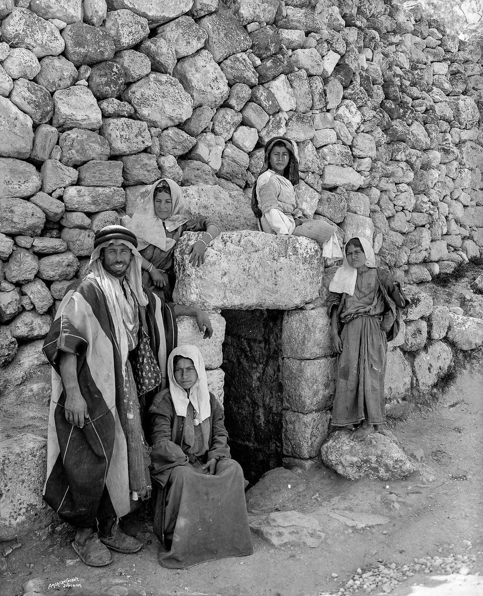Black-and-white photograph of a Bedouin family posed in front of an ancient stone structure. The group includes three women, a man, and a young girl, all wearing traditional clothing with head coverings. They stand or sit near a small arched doorway set into a dry-stone wall, likely in the Sinai or Jerusalem region, early 20th century.