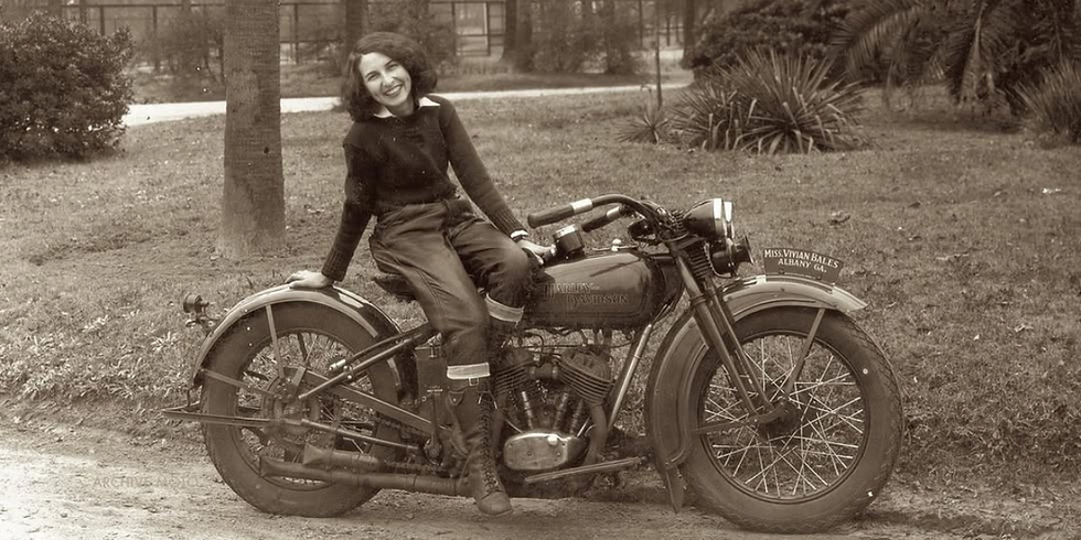 Vivian Bales smiling, sits on a vintage Harley-Davidson motorcycle in a park. Sign reads "Miss Vivian Bales, Albany GA." Sepia tones.