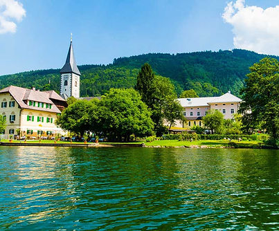 Dorf am See im Sommer mit Kirche und bewaldeten Bergen