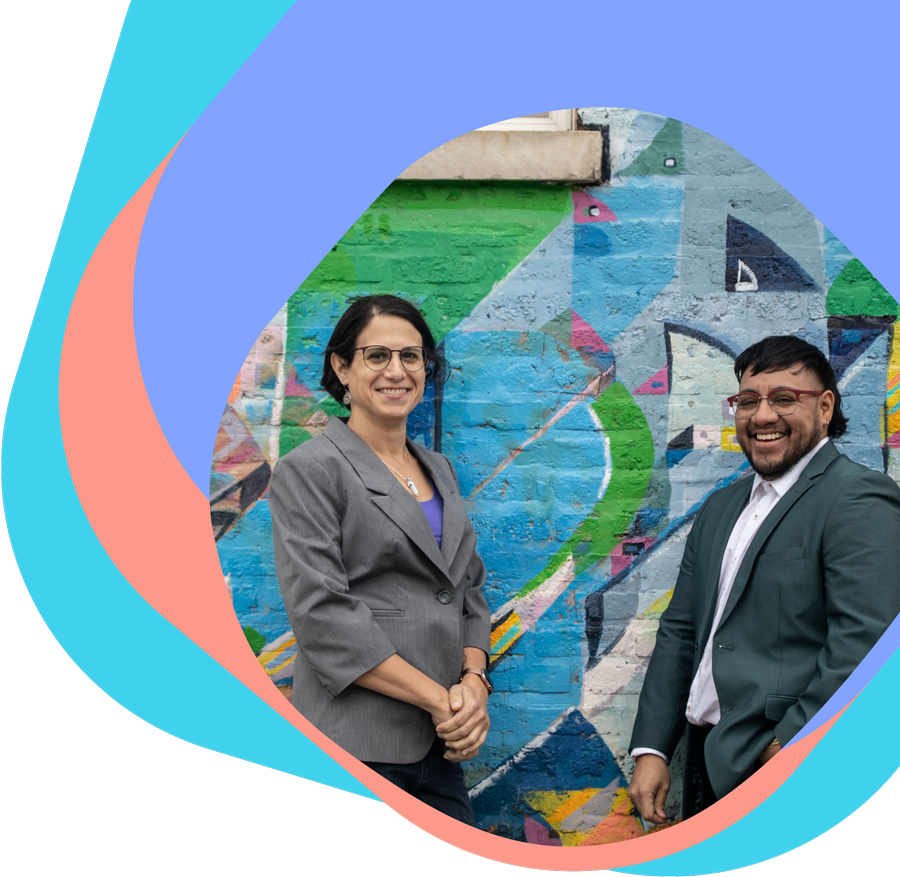 Photograph of Rebecca Kling and Crispin Torres smiling and leaning against a colorfully-painted brick wall
