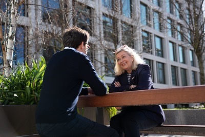 Helen Miles sitting on a bench speaking to another person. They are in front of an office block.
