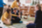 children working with montessori material on the floor of a classroom accross from their teacher