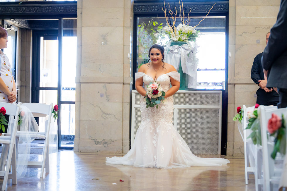 Bride walks down aisle in elegant mermaid wedding gown holding pink rose bouquet at indoor ceremony venue