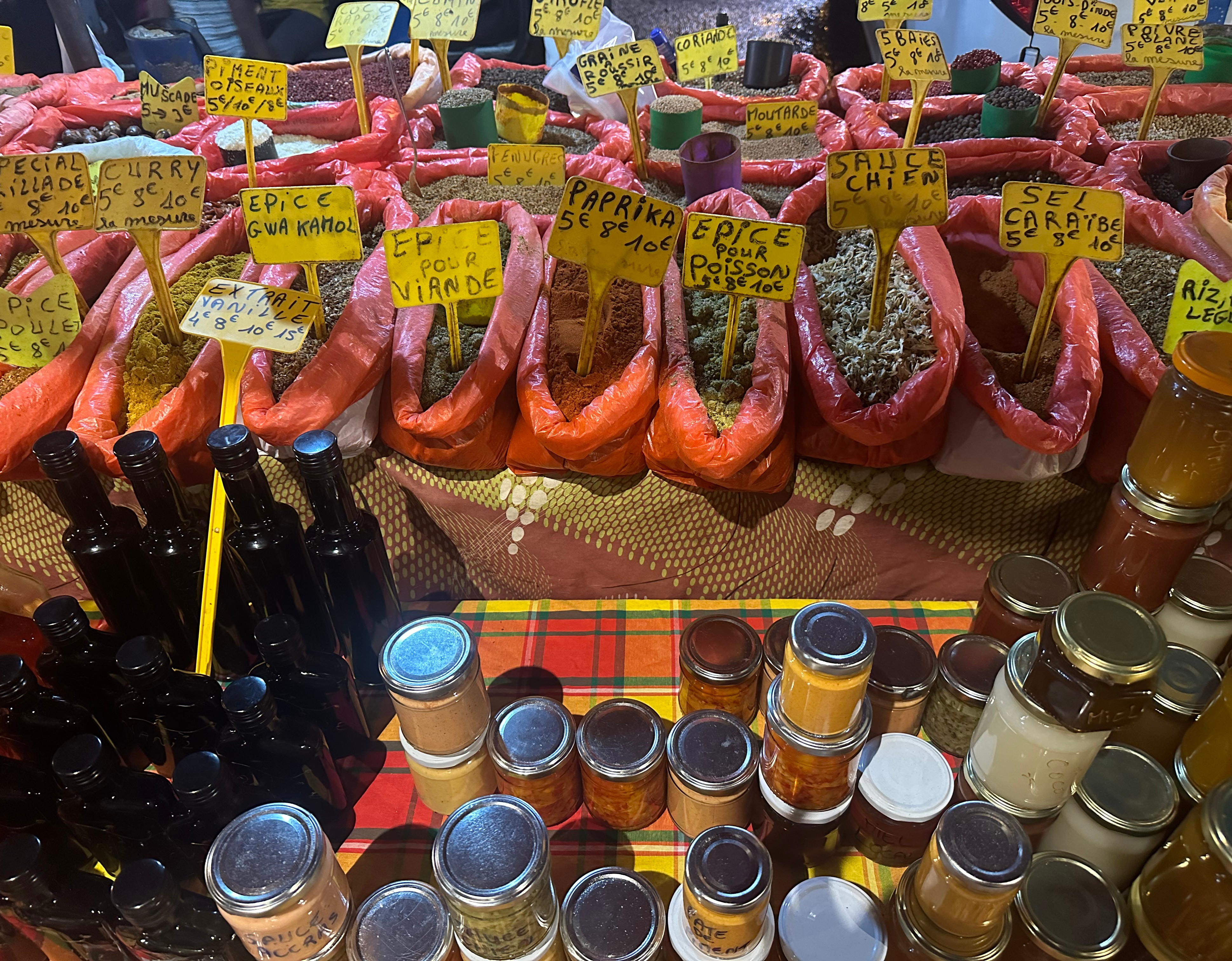Étale de marché en Guadeloupe avec des épices colorées, des bocaux de confitures et condiments et des bouteilles d'essence de vanille.